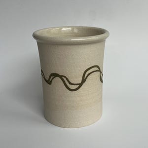 May include: A beige ceramic tumbler with a wavy brown design. The cylindrical cup has a slightly flared rim and a smooth, unglazed surface. The brown design wraps around the body of the cup.