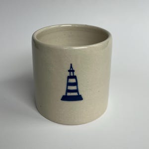 May include: A cylindrical, off-white ceramic mug with a blue lighthouse design. The mug has a smooth, unglazed finish and a simple, minimalist design. The lighthouse graphic is centered on the front of the mug.