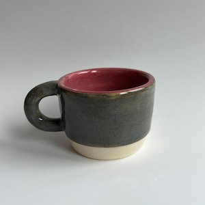 May include: A small ceramic mug with a dark gray exterior, a red interior, and a cream-colored base. The mug has a rounded handle and is likely handmade. The mug is suitable for coffee or tea.