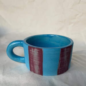 Puede incluir: Una pequeña taza de cerámica hecha a mano, turquesa brillante por fuera y por dentro. Dos rayas verticales granates están pintadas en el exterior. La taza tiene un asa redondeada y es adecuada para bebidas calientes.