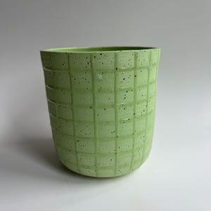Puede incluir: Un jarrón de cerámica verde claro con un patrón de rejilla texturizado. El jarrón tiene forma cilíndrica y está salpicado de pequeños puntos negros. El interior es de color verde claro liso.
