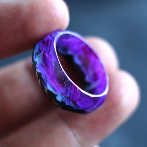 May include: A vibrant purple ring with swirling patterns of blue and white, resembling a galaxy. The ring is held in a hand, showcasing its unique design and color variations. The ring's interior is a lighter shade of purple.