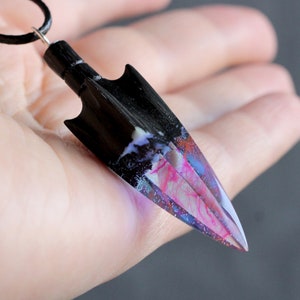 May include: A black arrowhead pendant with a colorful, galaxy-like resin inlay. The pendant is attached to a black cord.