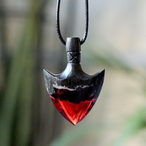May include: A handcrafted pendant necklace featuring a unique arrowhead design. The pendant combines dark wood with a vibrant red resin, creating a striking contrast. The necklace hangs from a black cord, offering a bold, artistic statement.