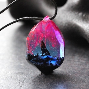 May include: A dark grey wolf silhouette is encased in a clear resin pendant, with a pink and purple aurora borealis effect in the background. The pendant is attached to a black cord.