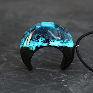 May include: A crescent moon-shaped pendant with a black wood base and a blue resin top. The resin is filled with white and blue swirls and sparkles.
