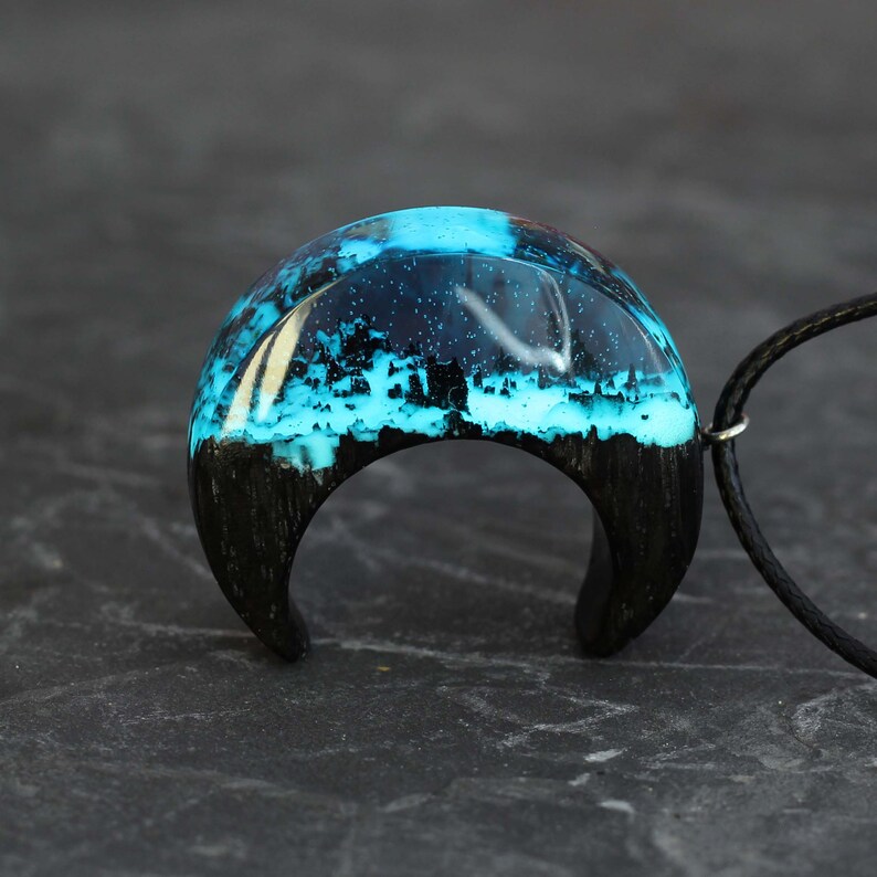 May include: A crescent moon-shaped pendant with a black wood base and a blue resin top. The resin is filled with white and blue swirls and sparkles.