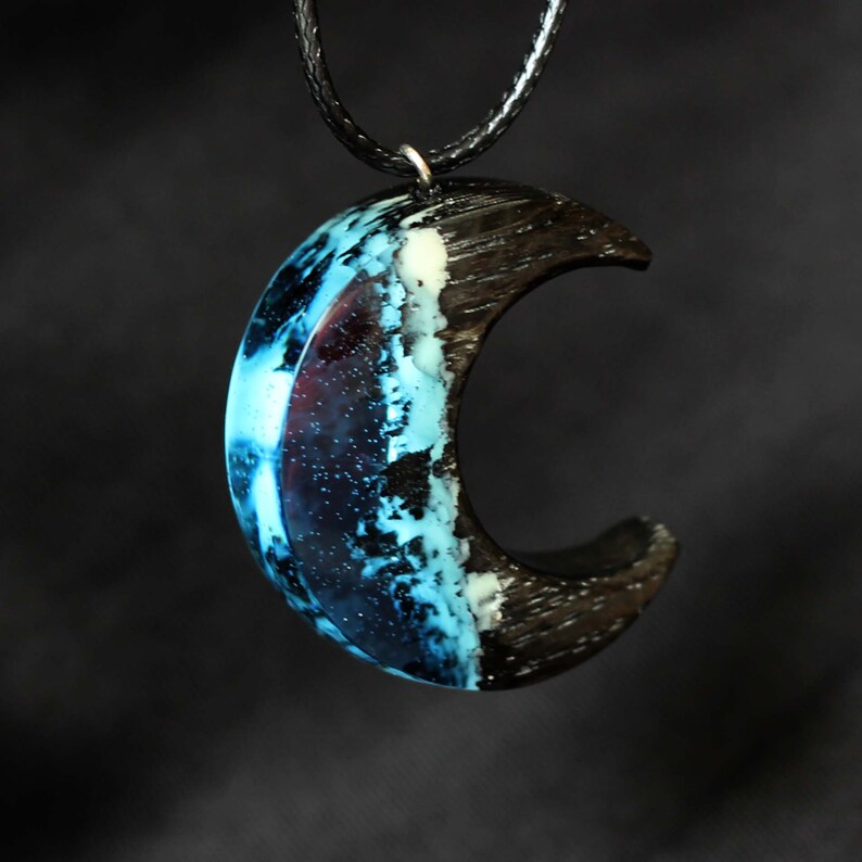 May include: A crescent moon pendant necklace made of black wood and blue resin. The resin has a swirling, galaxy-like pattern with white and black accents.