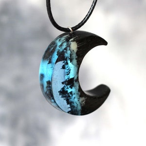 May include: A crescent moon pendant necklace made of black wood and blue resin. The resin has a swirling pattern that resembles a night sky with stars.