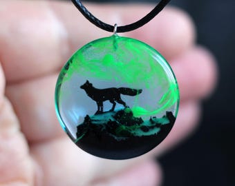 May include: A round pendant necklace with a black cord. The pendant features a black silhouette of a wolf standing on a mountain range with a green and blue aurora borealis in the background.