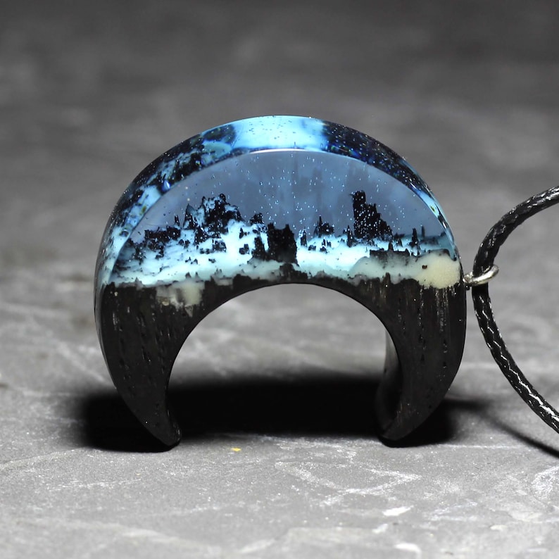 May include: A crescent moon shaped pendant with a black wood base and a clear resin top. The resin contains a cityscape scene with white buildings and a blue sky.