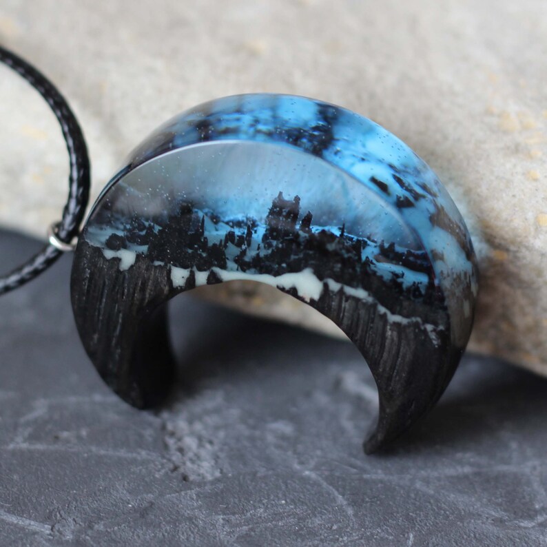 May include: A crescent moon shaped pendant necklace with a black wood base and a clear resin top. The resin contains a blue and white cityscape scene.