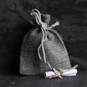 May include: A small gray burlap sack with a white paper note tied to it with twine. The sack is tied with a gray string.