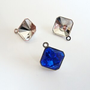 May include: Three square-shaped crystal charms with metal loops. Two charms are clear and one is blue.