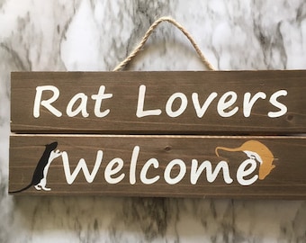 May include: A wooden sign with the text "Rat Lovers Welcome" in white paint. The sign is brown and has two rats painted on it, one black and one orange.