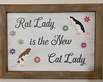 May include: A framed sign with the text "Rat Lady is the New Cat Lady" in black cursive font. The sign is decorated with colorful paper flowers and two paper rat cutouts.