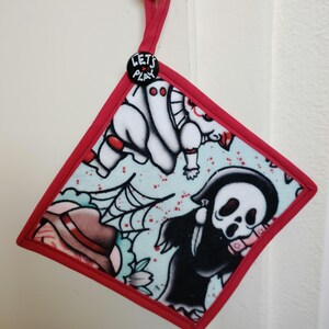 May include: A diamond-shaped pot holder with a red border and a red loop for hanging. The fabric features a light blue background with tattoo-style illustrations of a ghost, a spider web, and a grim reaper. A black button with the text "LET'S PLAY" is attached.