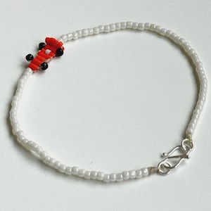 May include: White beaded bracelet with a red and black airplane charm. The bracelet has a silver clasp. The beads are small and round. The airplane charm is made of small beads.