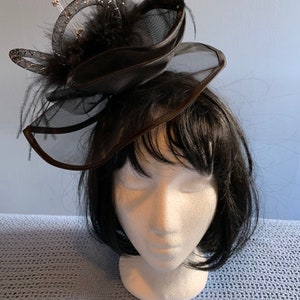 Könnte beinhalten: Ein schwarzer Fascinator-Hut mit einem mehrschichtigen Design und einem Federschmuck. Der Hut besteht aus einer Kombination aus Netz- und Satin-Stoff.
