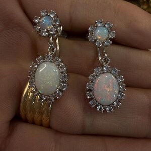 May include: A pair of opal drop earrings. Each earring features a large, oval opal surrounded by a halo of small, clear gemstones. The earrings have a floral design at the top and are suspended from a small hook.