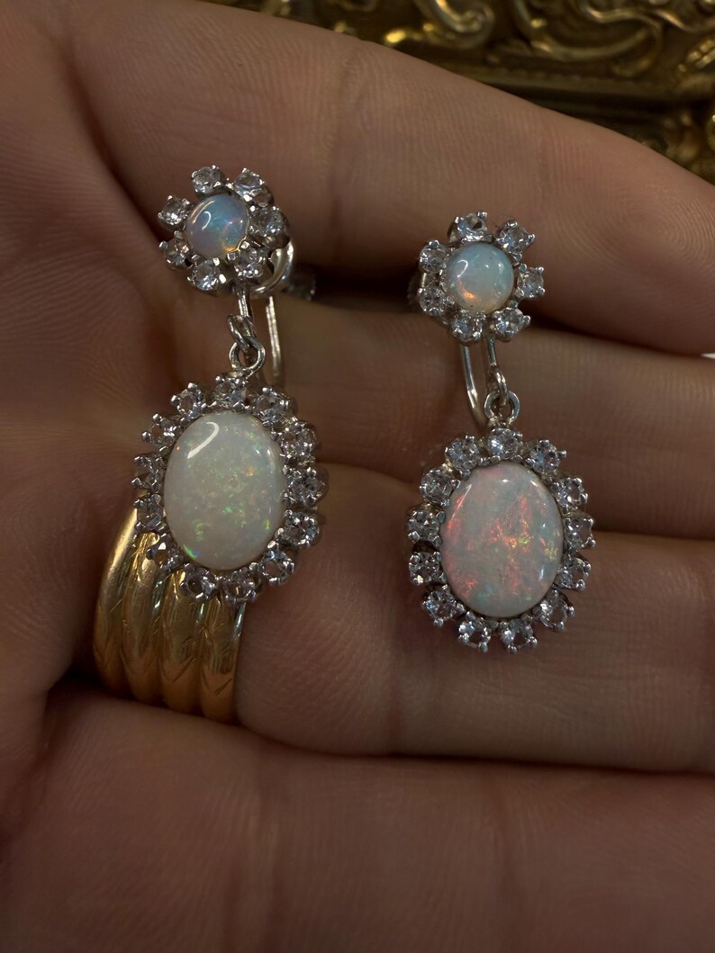 May include: A pair of opal drop earrings. Each earring features a large, oval opal surrounded by a halo of small, clear gemstones. The earrings have a floral design at the top and are suspended from a small hook.