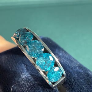 May include: A silver ring featuring five round, faceted, light blue gemstones. The gemstones are set in a channel setting, creating a continuous line of color. The ring is displayed on a dark blue velvet surface.