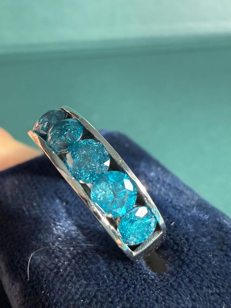 May include: A silver ring featuring five round, faceted, light blue gemstones. The gemstones are set in a channel setting, creating a continuous line of color. The ring is displayed on a dark blue velvet surface.