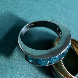 May include: A silver ring with a row of blue gemstones and a gold ring on a teal surface. The silver ring has a polished finish, and the gemstones are a vibrant blue color. The gold ring is smooth and shiny.