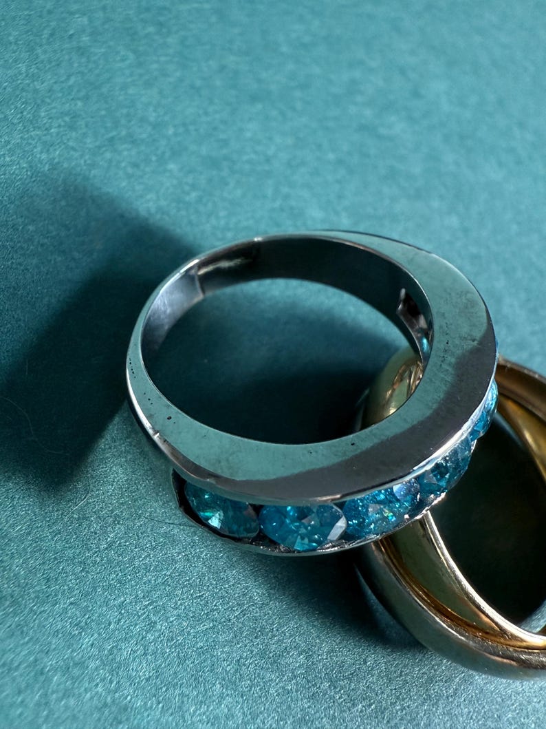 May include: A silver ring with a row of blue gemstones and a gold ring on a teal surface. The silver ring has a polished finish, and the gemstones are a vibrant blue color. The gold ring is smooth and shiny.