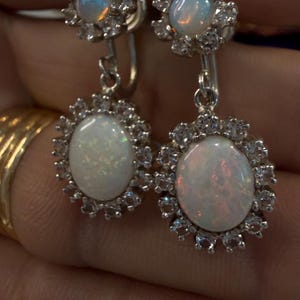 May include: A pair of silver-toned drop earrings featuring oval opals surrounded by a halo of small, clear gemstones. The earrings have a floral design at the top and are suspended from a lever back closure.