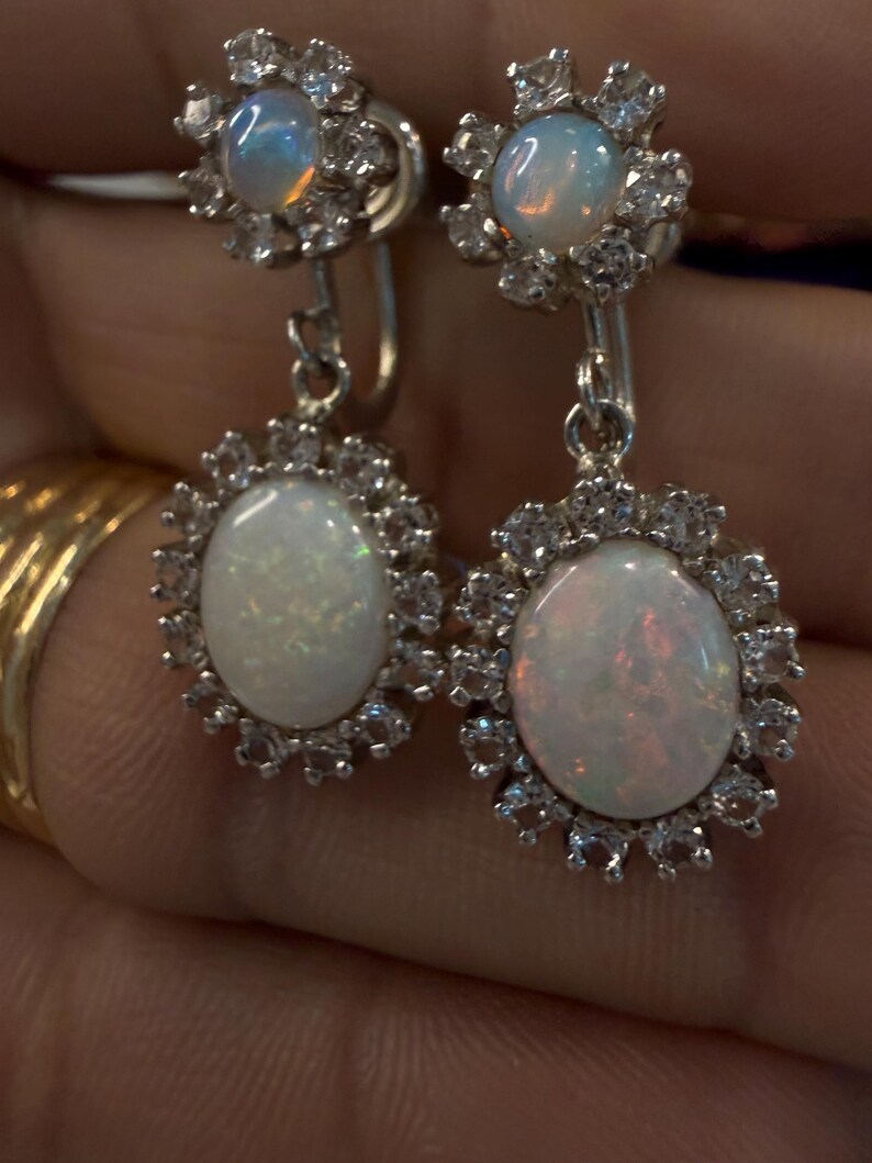 May include: A pair of silver-toned drop earrings featuring oval opals surrounded by a halo of small, clear gemstones. The earrings have a floral design at the top and are suspended from a lever back closure.