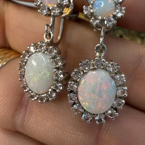 May include: A pair of silver-toned earrings featuring oval opals surrounded by small, clear gemstones. The opals have a milky white base with flashes of color. The earrings have a floral design and a dangling element.