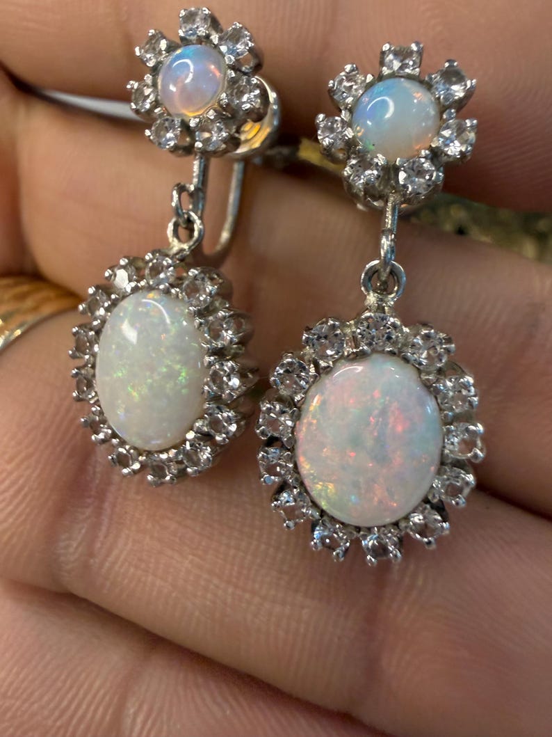 May include: A pair of silver-toned earrings featuring oval opals surrounded by small, clear gemstones. The opals have a milky white base with flashes of color. The earrings have a floral design and a dangling element.