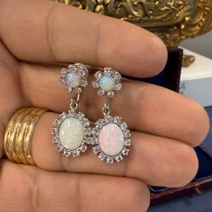 May include: A pair of silver earrings featuring oval opal gemstones surrounded by a halo of small, clear crystals. Each earring has a smaller opal at the top, connected by a silver chain. The earrings are displayed against a neutral background.