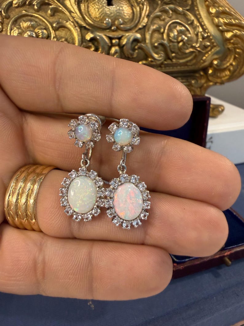 May include: A pair of silver earrings featuring oval opal gemstones surrounded by a halo of small, clear crystals. Each earring has a smaller opal at the top, connected by a silver chain. The earrings are displayed against a neutral background.