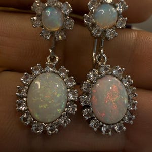 May include: A pair of silver drop earrings featuring oval opals surrounded by a halo of small, clear gemstones. The earrings have a floral design at the top and a matching halo around the opal drops. The opals display iridescent colors.