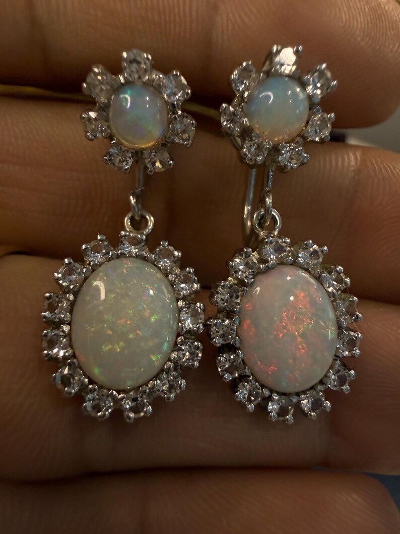 May include: A pair of silver drop earrings featuring oval opals surrounded by a halo of small, clear gemstones. The earrings have a floral design at the top and a matching halo around the opal drops. The opals display iridescent colors.
