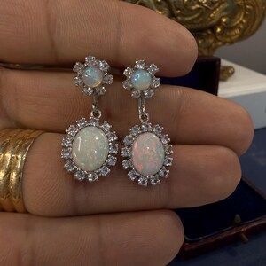 May include: A pair of silver-toned drop earrings. Each earring features an oval opal stone surrounded by small, clear gemstones. The top of each earring has a smaller opal stone surrounded by clear gemstones.