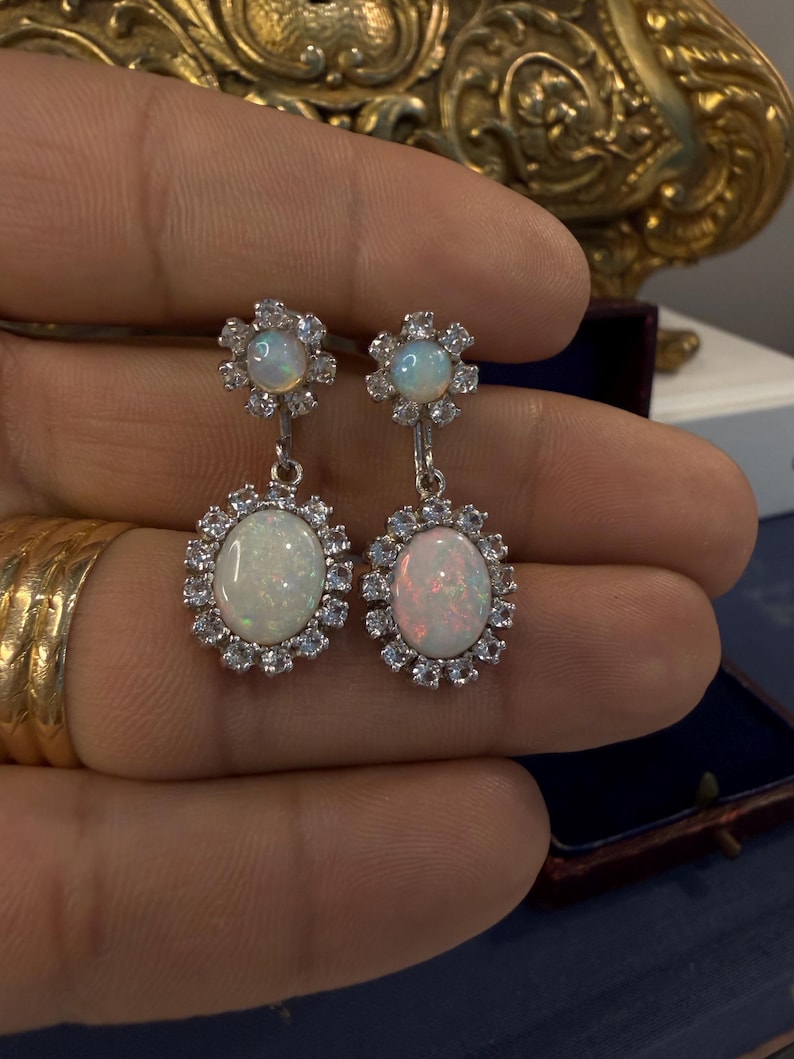 May include: A pair of silver-toned drop earrings. Each earring features an oval opal stone surrounded by small, clear gemstones. The top of each earring has a smaller opal stone surrounded by clear gemstones.