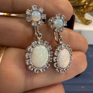 May include: A pair of silver earrings featuring oval opal gemstones surrounded by small, sparkling clear stones. The earrings have a floral design at the top and a larger oval opal at the bottom, creating a layered effect. The opals have a milky white color.