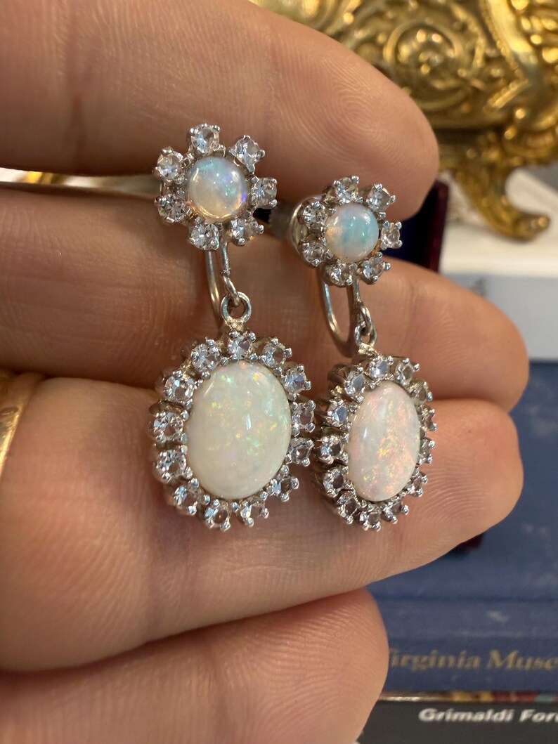 May include: A pair of silver earrings featuring oval opal gemstones surrounded by small, sparkling clear stones. The earrings have a floral design at the top and a larger oval opal at the bottom, creating a layered effect. The opals have a milky white color.