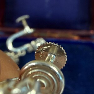May include: Close-up of a gold-colored earring screw back with a textured edge and a threaded post. The earring is marked "14K". The background is a blurred blue.
