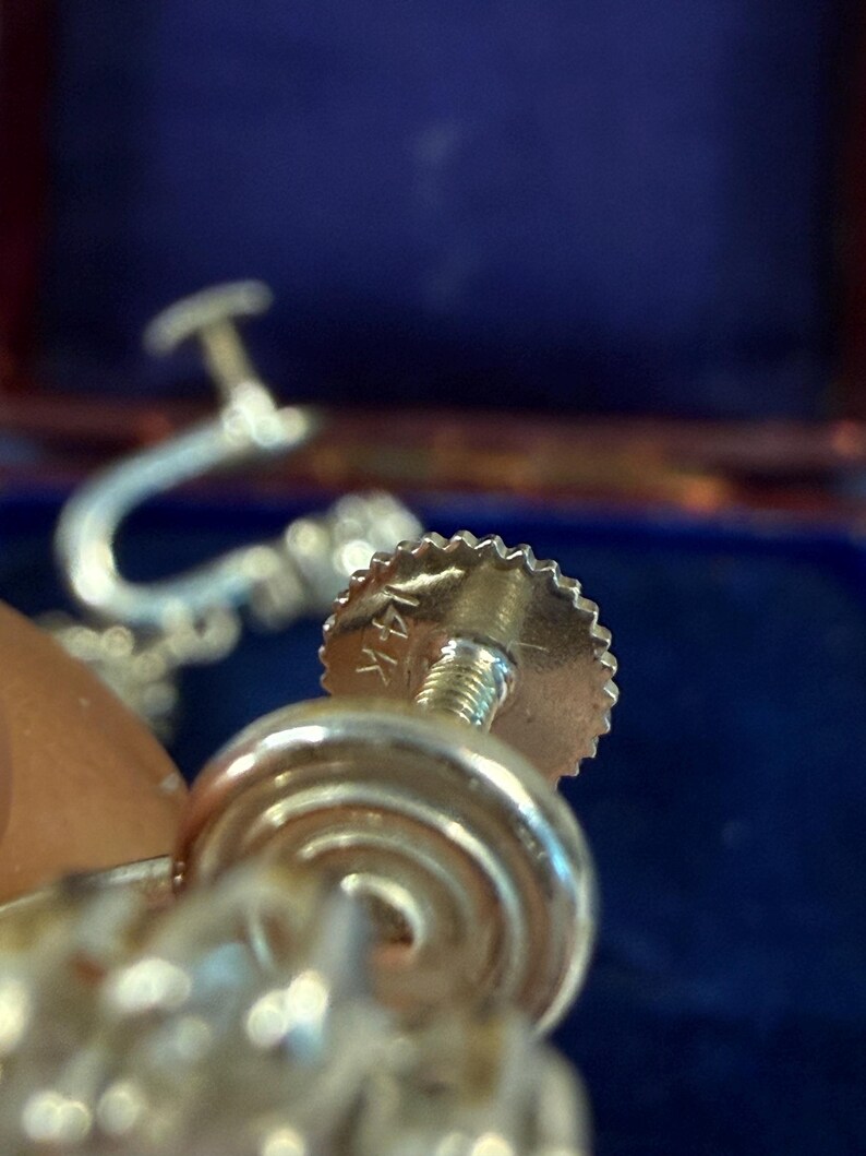 May include: Close-up of a gold-colored earring screw back with a textured edge and a threaded post. The earring is marked "14K". The background is a blurred blue.