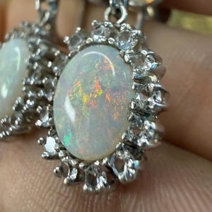 May include: A pair of silver-toned earrings featuring oval-shaped opals with iridescent flashes of color. Each opal is surrounded by a halo of smaller, clear gemstones. The earrings have a classic, elegant design.