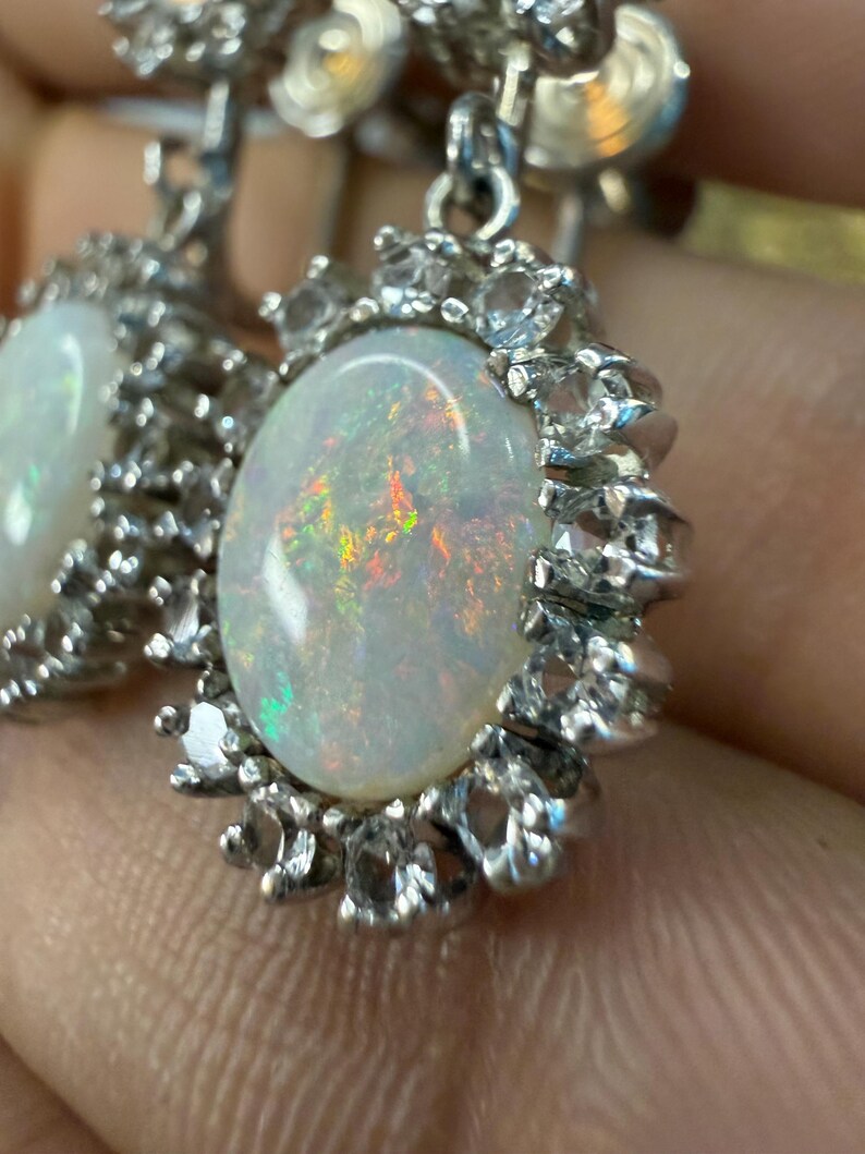 May include: A pair of silver-toned earrings featuring oval-shaped opals with iridescent flashes of color. Each opal is surrounded by a halo of smaller, clear gemstones. The earrings have a classic, elegant design.