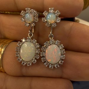 May include: A pair of silver earrings featuring opalescent oval-shaped gemstones, each surrounded by a halo of small, clear stones. The earrings have a floral design at the top and a dangling oval stone at the bottom.