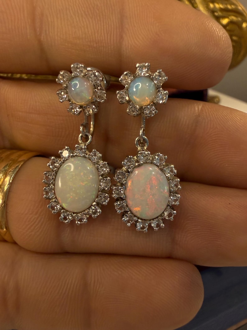 May include: A pair of silver earrings featuring opalescent oval-shaped gemstones, each surrounded by a halo of small, clear stones. The earrings have a floral design at the top and a dangling oval stone at the bottom.
