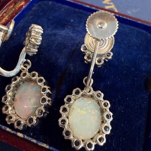 May include: A pair of vintage silver earrings with oval opal gemstones. Each opal is surrounded by a halo of small, clear gemstones. The earrings are displayed in a blue velvet jewelry box.