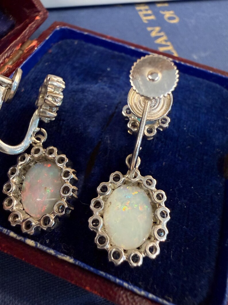 May include: A pair of vintage silver earrings with oval opal gemstones. Each opal is surrounded by a halo of small, clear gemstones. The earrings are displayed in a blue velvet jewelry box.