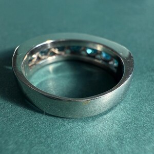 May include: A silver ring with a row of small, bright blue gemstones set into the band. The ring is shown on a teal-colored surface, with the focus on the gemstones and the polished metal.